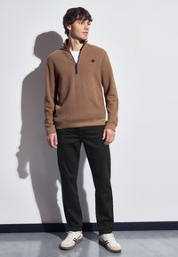 Joven de pie contra una pared blanca, vistiendo un suéter marrón con cremallera, pantalones negros y zapatillas blancas, mirando hacia un lado.