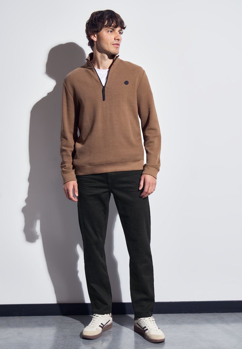 Joven de pie contra una pared blanca, vistiendo un suéter marrón con cremallera, pantalones negros y zapatillas blancas, mirando hacia un lado.