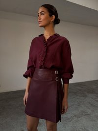 Maroon blouse with ruffled collar and buttons, paired with a matching leather skirt featuring buckle accents and a wrapped design.