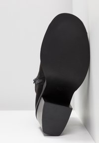 Semelle de chaussure à talon noire tournée vers l'avant, montrant un dessous texturé et un talon carré, positionnée contre un fond blanc et un coin de mur.