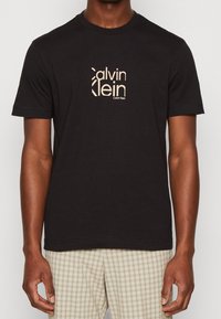 Svart bomullströja med en central beige Calvin Klein-logotyp. Den har korta ärmar och rund halsringning med en rak fåll.