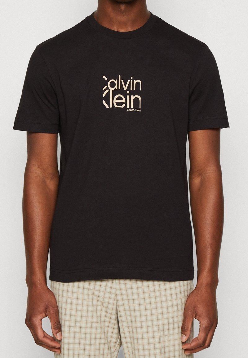 Svart bomullströja med en central beige Calvin Klein-logotyp. Den har korta ärmar och rund halsringning med en rak fåll.