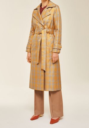 Vrouw draagt een lange beige en geel geruite trenchcoat met riem, bruine wijde broekspijpen en roestrode puntige schoenen tegen een effen achtergrond.