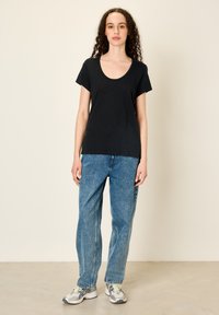 Jaunas asmuo su garbanotais plaukais, dėvintis juodą trumpomis rankovėmis marškinėlį, mėlynas džinsus ir baltus sportbačius, stovintis prie smėlio spalvos sienos.