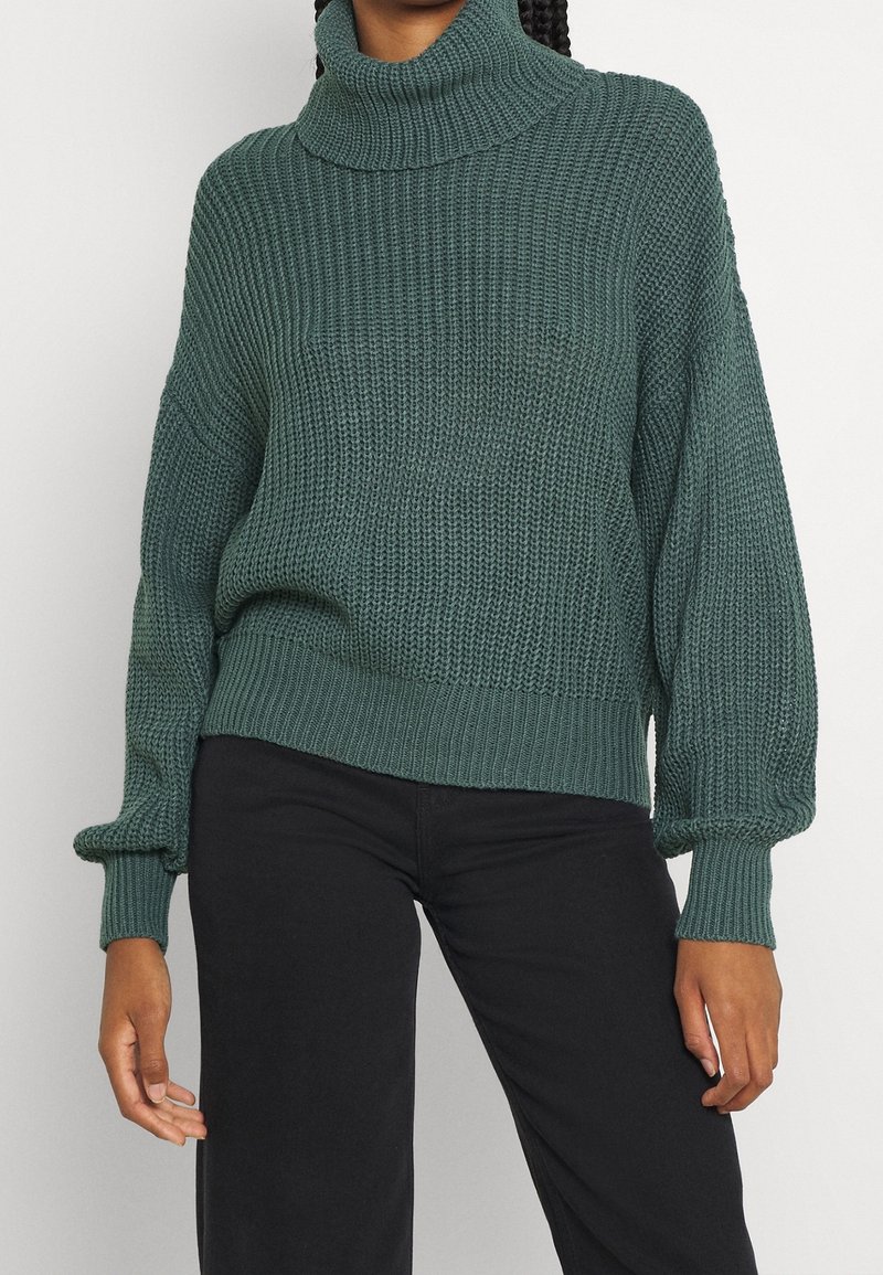 Femme portant un pull à col roulé en maille épaisse vert foncé avec des manches bouffantes et un pantalon noir, sur un fond uni.
