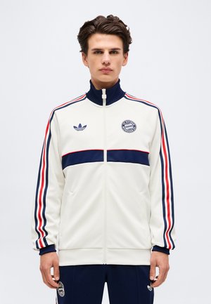 Giovane uomo che indossa una giacca sportiva bianca con zip con strisce blu navy e rosse, loghi FC Bayern Monaco e Adidas, in piedi davanti a uno sfondo semplice.