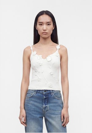 Femme aux longs cheveux noirs portant un haut sans manches blanc brodé de fleurs et un jean bleu, debout devant un fond uni.