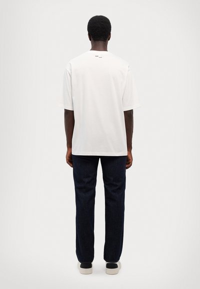 White short-sleeve t-shirt with loose fit, featuring a minimal design and a small black logo detail on the back, paired with dark jeans.