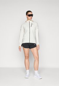 Letvægts cream jakke med en sort lynlås og hætte med orange detaljer, parret med sorte shorts. Modellen bærer solbriller og hvide sneakers.