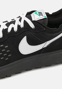 Sapatilha preta com parte superior em camurça e malha, logótipo Nike swoosh branco, atacadores brancos, detalhes texturizados na lateral e sola de borracha preta.