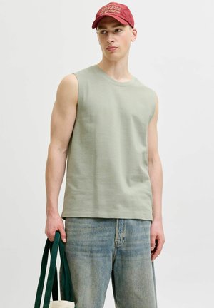 Jeune homme portant un débardeur vert poussiéreux, un jean bleu délavé et une casquette rouge, tenant un sac fourre-tout avec des poignées vertes.