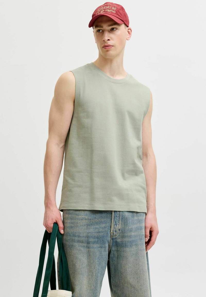 Jeune homme portant un débardeur vert poussiéreux, un jean bleu délavé et une casquette rouge, tenant un sac fourre-tout avec des poignées vertes.