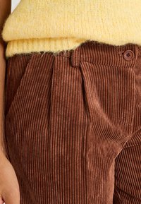 Un pantalon en velours côtelé marron présente des côtes verticales proéminentes, un bouton à la taille et des plis amples à l'avant. Le tissu a une texture douce.