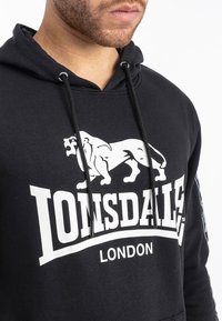 Black cotton hoodie with a white lion graphic and "Lonsdale London" text. Features drawstrings and a soft, textured finish.