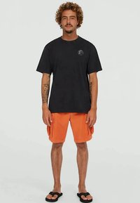 Hombre con cabello rizado que lleva una camiseta negra, pantalones cortos de carga naranjas, chanclas, de pie con los brazos relajados a los lados sobre un fondo sólido.