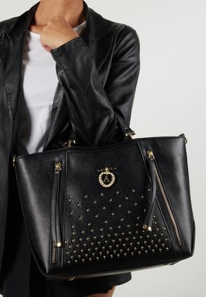 Sort læderhåndtaske med guld-nitter, en rund metalaccent og to lynlåslommer på siderne. Struktureret design med to top håndtag.