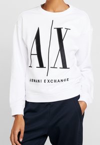 Felpa bianca con colletto a girocollo, caratterizzata da un grande logo nero "AX" e la scritta "ARMANI EXCHANGE" all'orlo. Tessuto morbido, vestibilità rilassata.