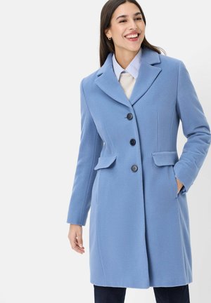 Lächelnde Frau, die einen blau geknöpften Mantel mit Klappentaschen, einen hellen Pullover und ein Hemd mit Kragen trägt, steht mit einer Hand in der Tasche.