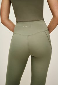 Aktív sportruházat leggings, tompa olivazöld színben, magas derékkal, sima textúrával és minimális logóval a hátsó részen.