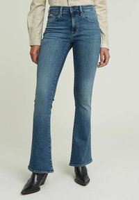 Uppsydda jeans i medelblå denim med en slät textur. Har en hög midja, framfickor och subtila sömdetaljer, kombinerat med svarta klackstövlar.