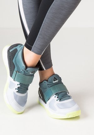 Træningssko med turkis mesh, hvide såler og CrossFit-mærkning, båret af en person i grå og sorte leggings med hvide striber.