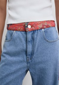 Jeans de mezclilla con lavado en azul claro, que cuentan con una vibrante cinturilla de patrón rojo y un botón a presión plateado. Diseño casual y ajustado con bolsillos frontales.