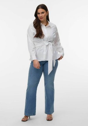 Kobieta o długich ciemnych włosach, ubrana w białą koszulę zapinaną na guziki, zawiązaną w talii, niebieskie dżinsy i sandały na obcasie, stojąca na białym tle.