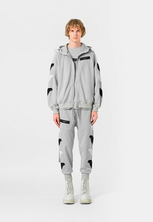 Joven de pie vistiendo una sudadera con capucha gris claro y pantalones jogger a juego con parches de alas en blanco y negro, y botas blancas con cordones.