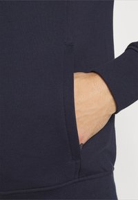Mano parzialmente dentro della tasca di un felpa blu navy con cerniera visibile su uno sfondo chiaro.