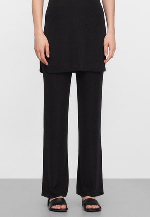 Dolna część ciała osoby noszącej czarne szerokie spodnie, dopasowaną długą tunikę oraz czarne klapki na palce, stojącej na szarej podłodze.