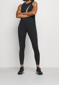 Femme debout, bras croisés, portant un débardeur de sport noir avec un motif blanc, un legging noir avec des bandes latérales et des baskets noires.