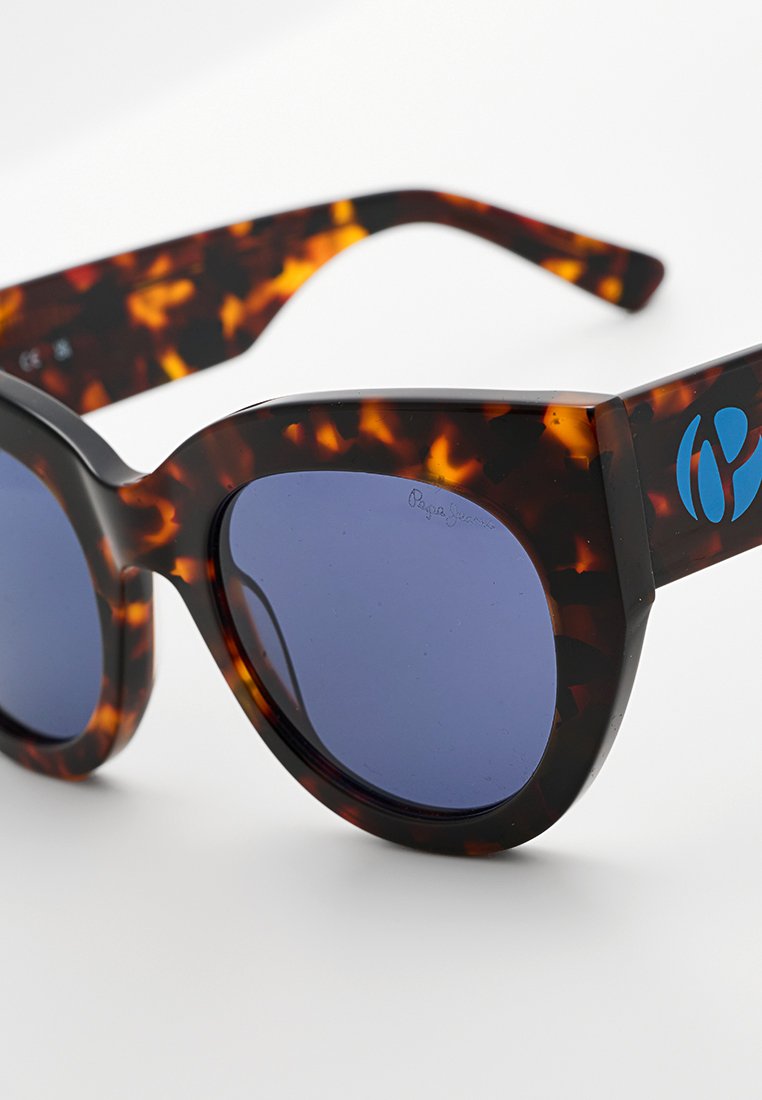Schildpatt-Cat-Eye-Sonnenbrille mit dunkelblauen Gläsern und einem kleinen blauen Logo am Bügel, auf einem weißen Hintergrund dargestellt.