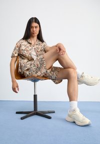 Camicia a maniche corte con bottoni e pantaloni corti in una stampa leopardata beige e marrone, abbinata a sneakers crema e calzini bianchi, seduto su una sedia di legno.
