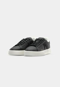Sorte lær sneakers med hvite såler, flate snørebånd, polstret tunge og en subtil logo detalj på siden. Jevn tekstur generelt.