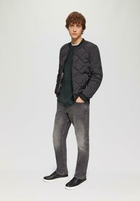 Gepolsterte schwarze Bomberjacke, strukturierter dunkelgrüner Strickpullover, graue Jeans, schwarze Sneakers mit weißen Sohlen; Model steht vor einem neutralen Hintergrund.