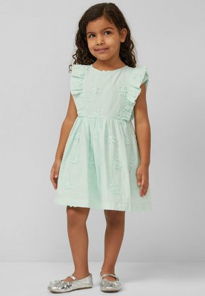 Junges Mädchen trägt ein hellblaues Kleid mit Rüschenärmeln und Blumenstickerei, steht vor einem schlichten grauen Hintergrund.