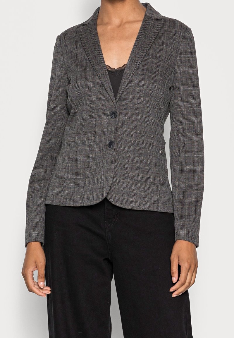 Vrouw in een grijs geruite blazer met twee zwarte knopen, over een zwarte top met kant en zwarte broek, staande tegen een effen achtergrond.