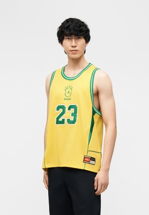 Jeune homme portant un maillot de basket-ball jaune du Brésil avec le numéro 23 et une bordure verte, debout devant un fond clair uni.