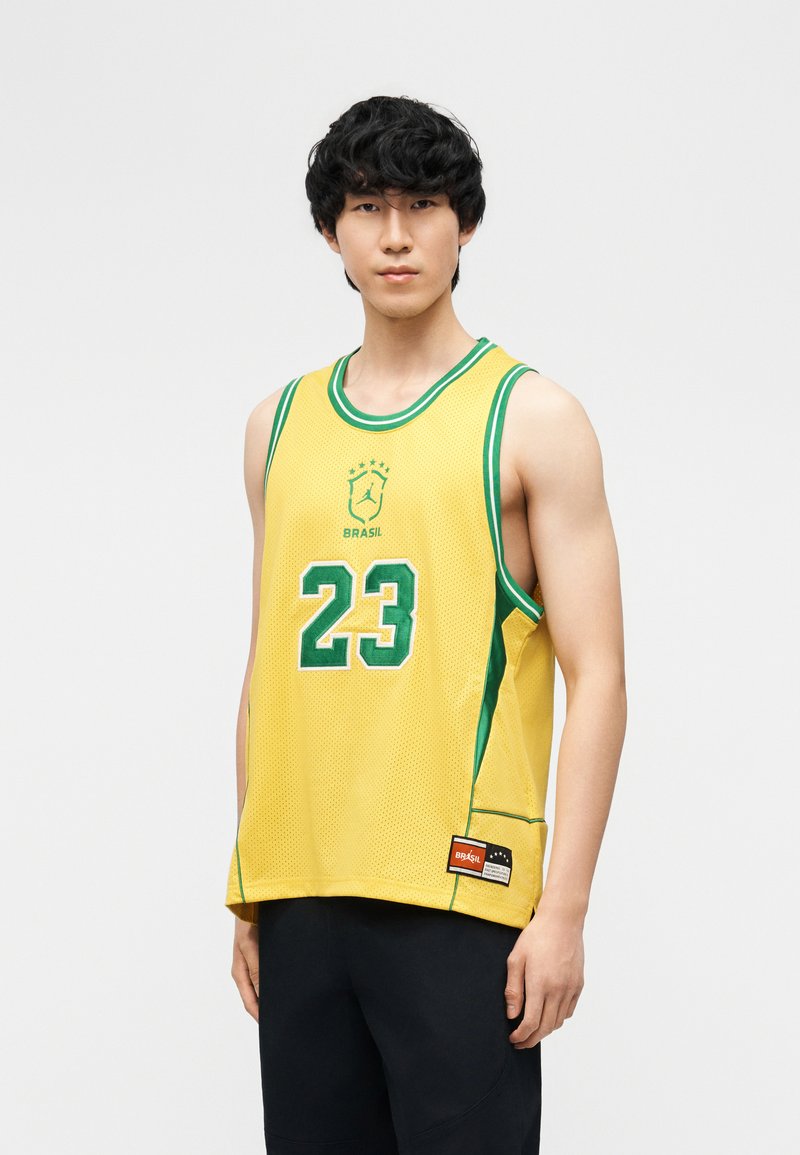 Νέος άνδρας που φοράει κίτρινη φανέλα μπάσκετ της Βραζιλίας με αριθμό 23 και πράσινη επένδυση, στέκεται μπροστά σε ένα απλό φωτεινό φόντο.