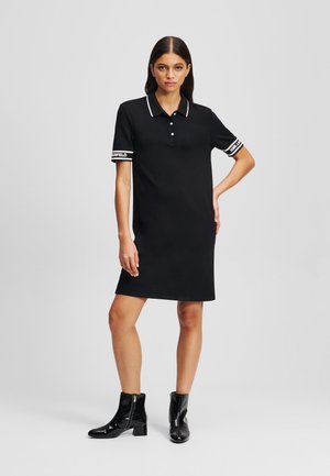 Abito polo nero con maniche corte, colletto bianco e dettagli a righe. Il materiale appare liscio e leggero. Abbinato a stivaletti neri lucidi alla caviglia.