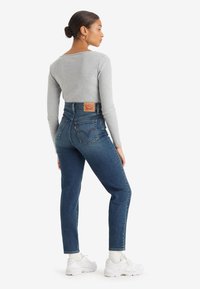 Jeans de ganga azul de cintura alta com um corte clássico, apresentando um patch de logo em pele nas costas, combinados com uma blusa justa de manga longa cinza.