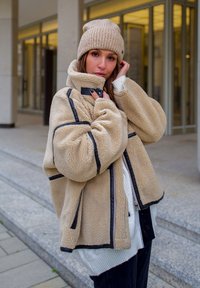 Beige fleece jacket with black trim, featuring a high collar and zip closure. Worn over a white sweater, complemented by a light beige knit beanie.