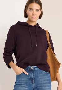 Sudadera morada con cordones y puños acanalados, combinada con jeans azules. Un bolso de hombro de ante color tostado completa el look.
