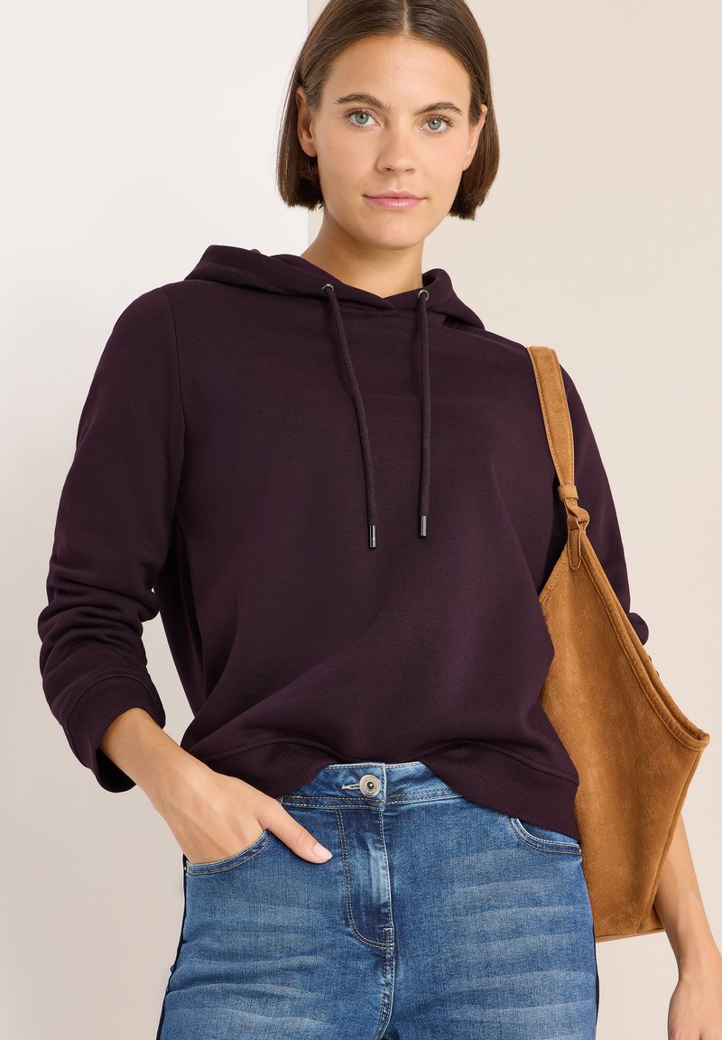 Sudadera morada con cordones y puños acanalados, combinada con jeans azules. Un bolso de hombro de ante color tostado completa el look.
