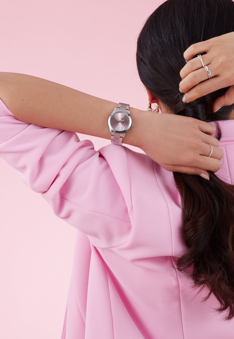 Montre en argent avec un cadran rond et un bracelet métallique, dotée d'un cadran rose, d'indices et d'une couronne simple. Portée sur un poignet devant un fond rose.