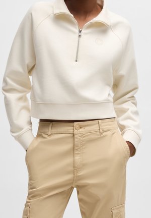 Pull court crème avec demi-fermeture éclair et pantalon beige taille haute avec poches avant et cargo, porté avec les mains dans les poches.