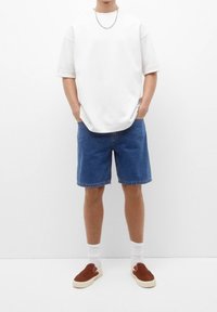 Camiseta blanca de gran tamaño, pantalones cortos de mezclilla, zapatos marrones de slip-on, calcetines blancos. Diseño simple, corte relajado, con mangas cortas y cuello redondo.