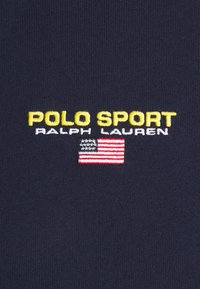 Dark navy fabric featuring "POLO SPORT" in yellow, "RALPH LAUREN" in white, and a small embroidered American flag in red, white, and blue.
