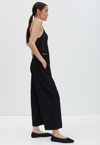 Vrouw met een lange paardenstaart, gekleed in een zwarte mouwloze top, wijde zwarte broek en zwarte flats, staat zijwaarts met haar handen in haar zakken.