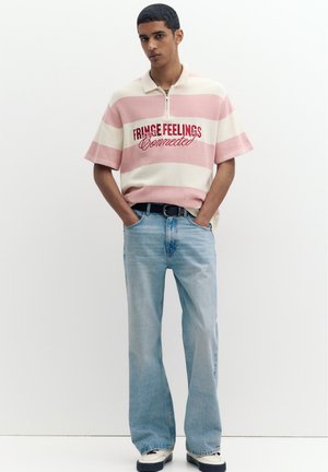 Junger Mann trägt ein pink-weiß gestreiftes Polo-Shirt mit dem Text „FRINGE FEELINGS Connected“, hellblaue Jeans und weiße Sneaker, Hände in den Taschen.